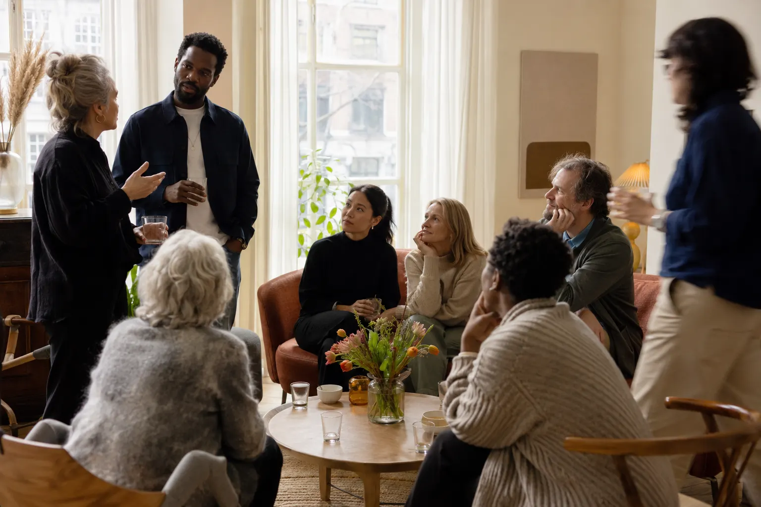 Een groep mensen in gesprek in een lichte huiskamer tijdens een bijeenkomst; een vrouw staat en spreekt, de anderen luisteren.